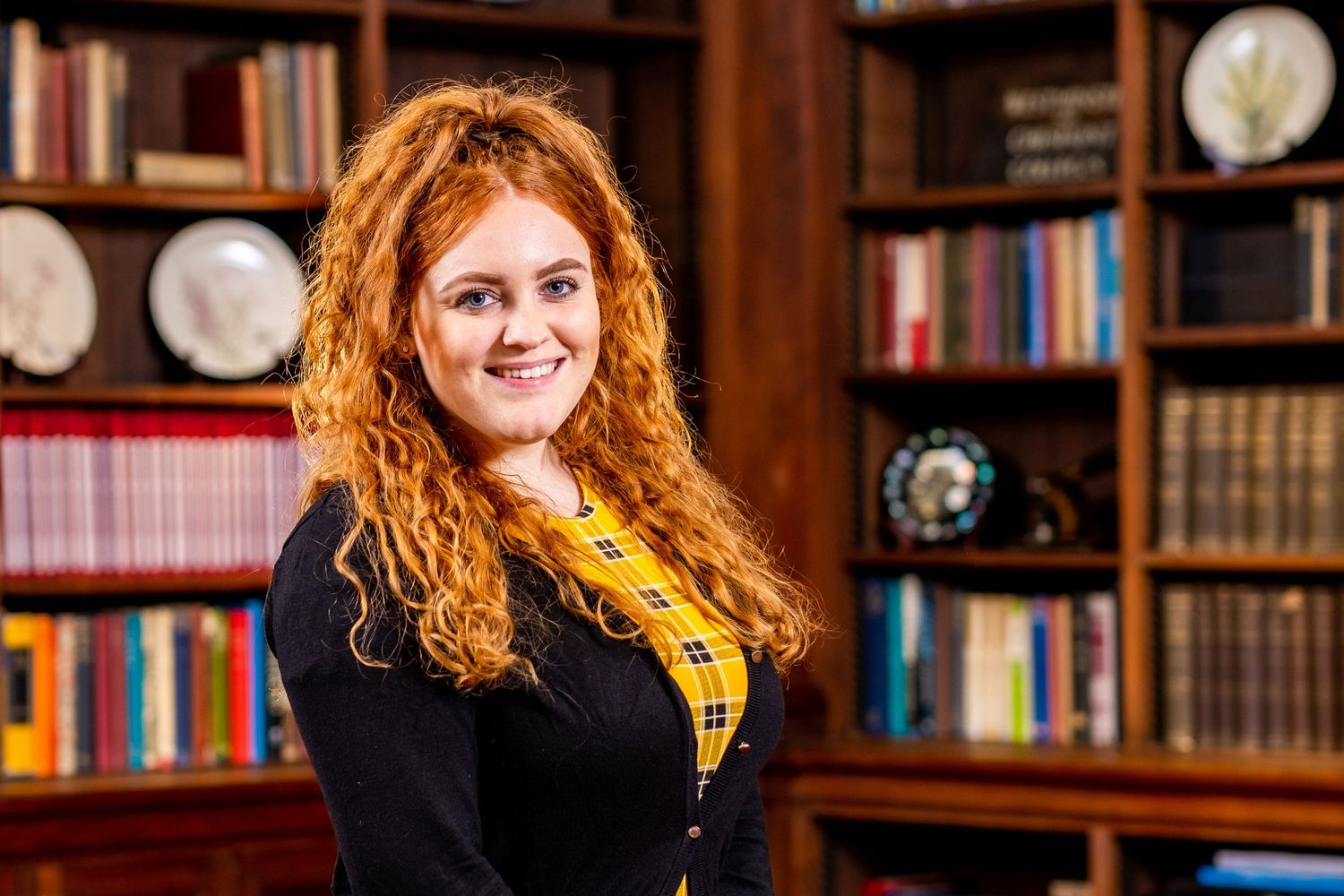 Woman with curly red hair wearing yellow scarf shown in library setting with bookshelves.