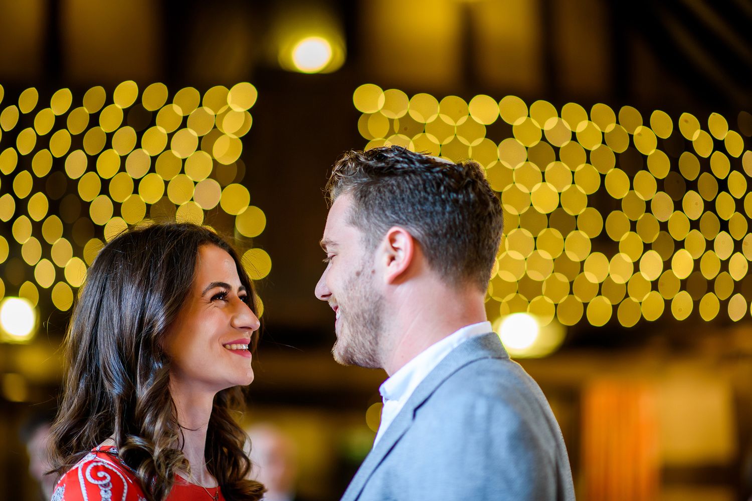 Romantic engagement photos under glowing string lights creating bokeh effect.