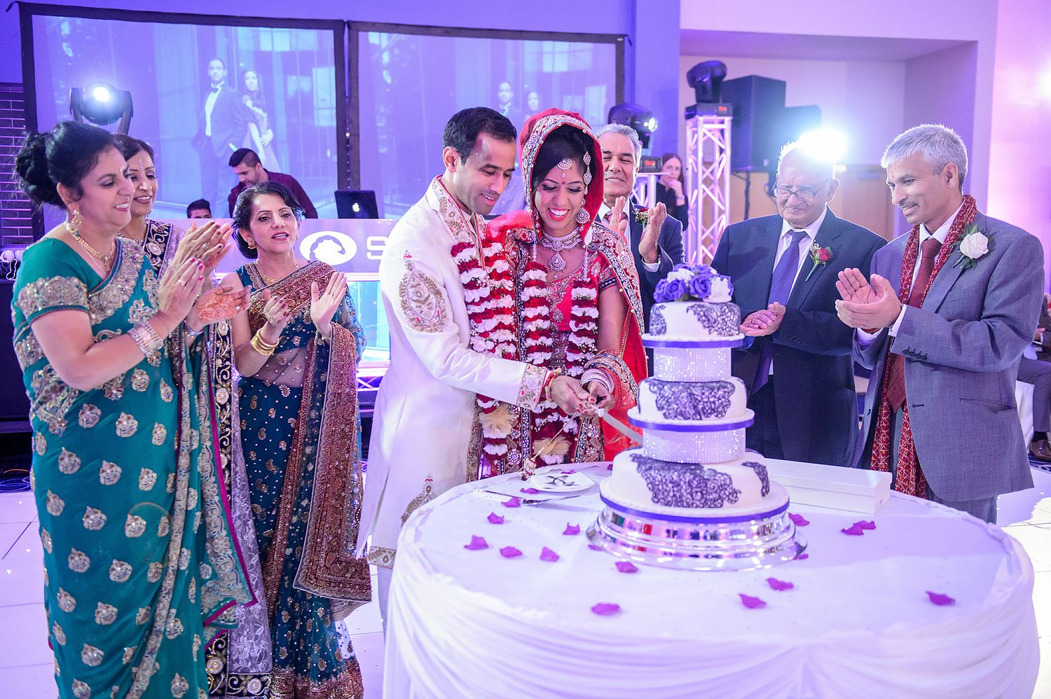 Wedding guests surround a beautiful purple-lit wedding cake during a traditional South Asian celebration.
