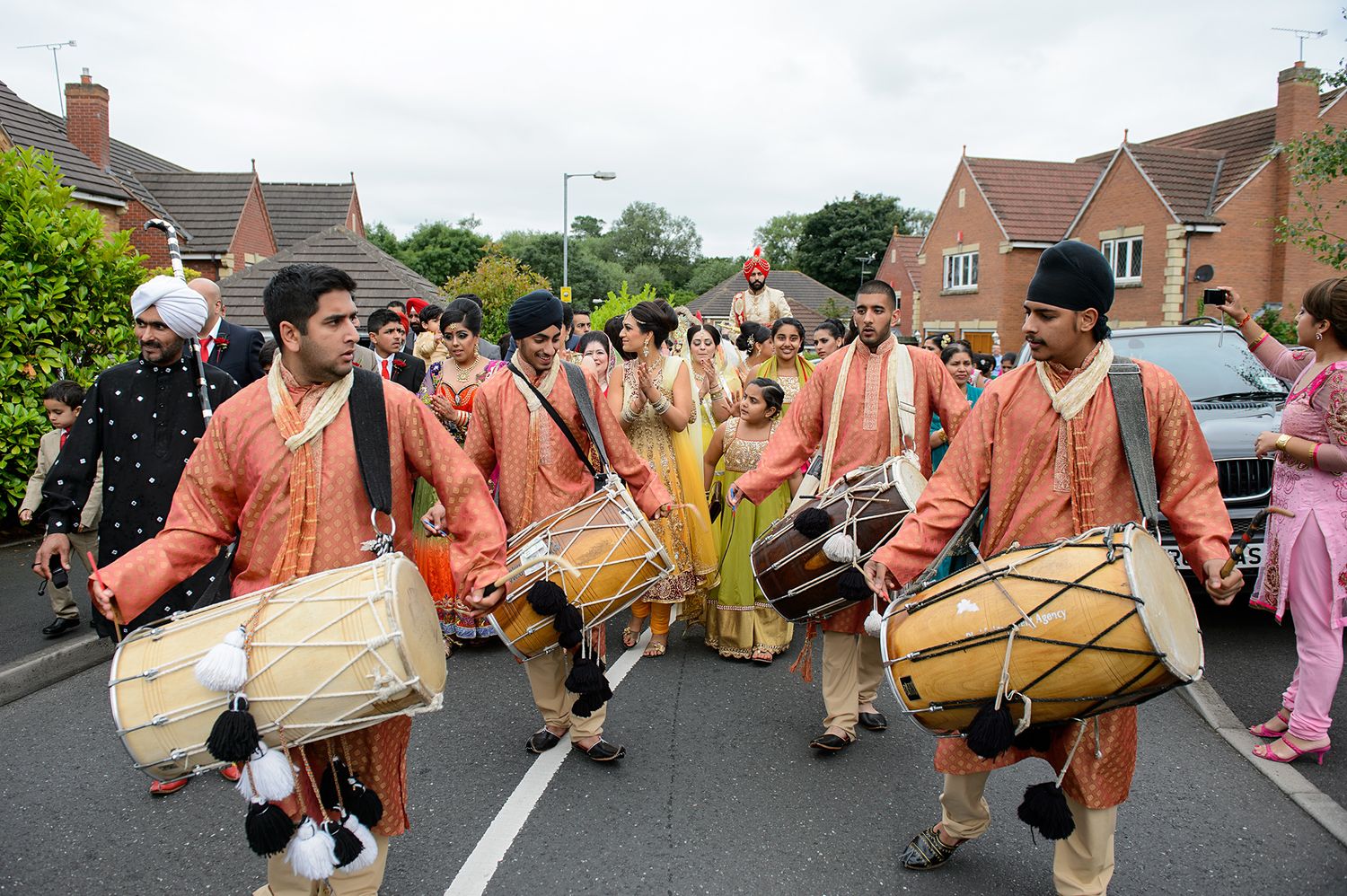 Traditional dhol drummers in peach outfits perform during an Indian wedding procession in a residential street.