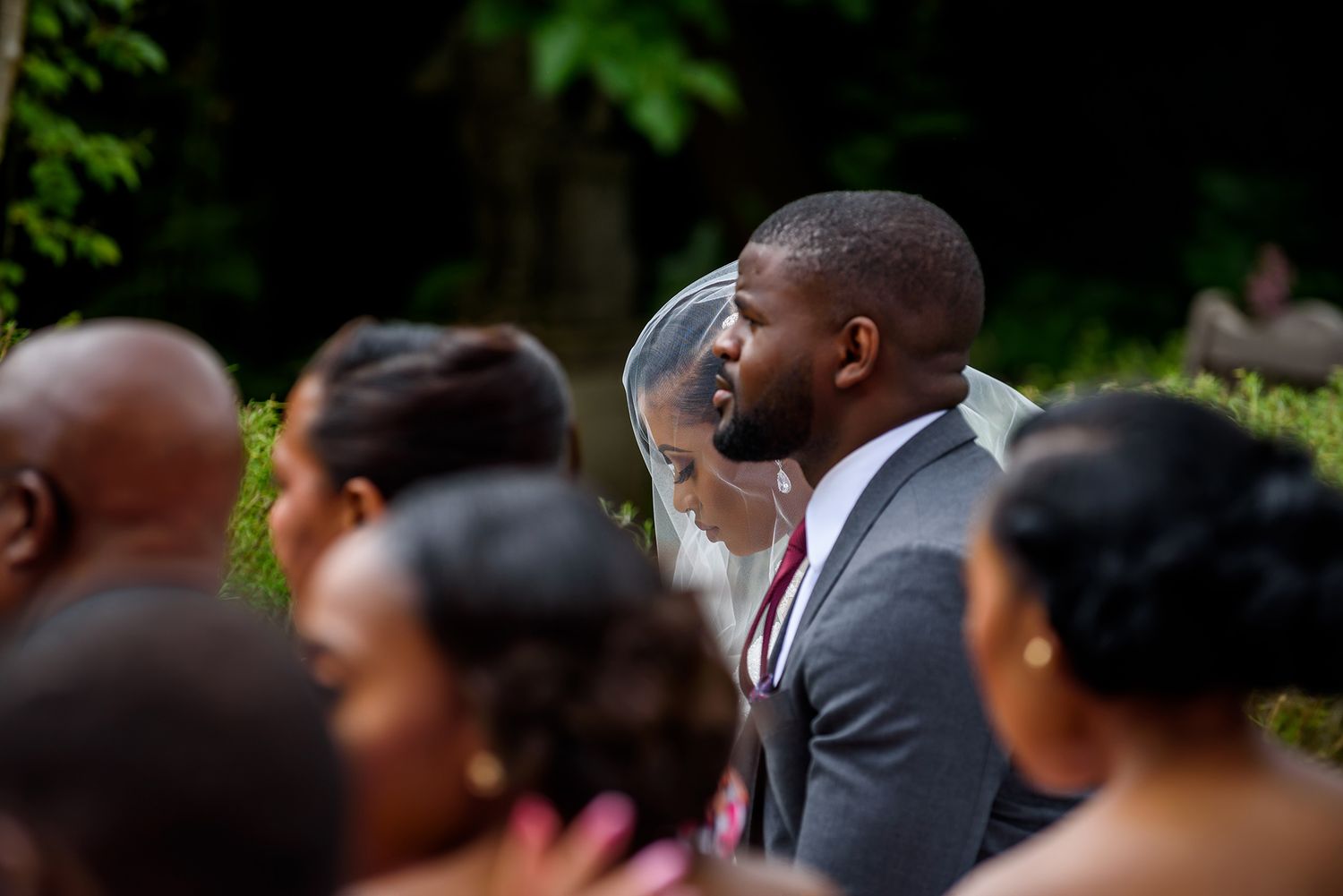 Wedding guests gather outdoors under green foliage during summer ceremony.