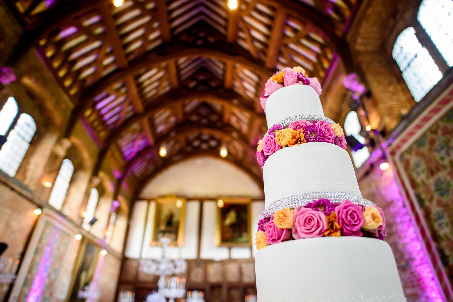 Elegant wedding cake with pink roses displayed in ornate wooden venue with vaulted ceiling.