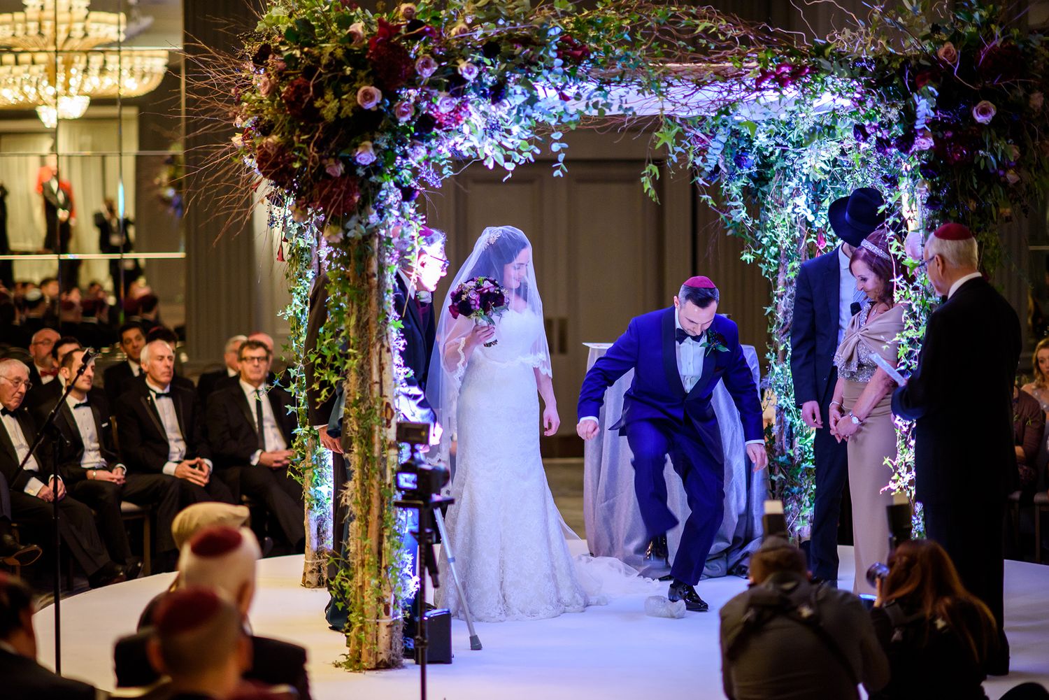 Wedding ceremony under illuminated floral arch with guests seated in formal attire.