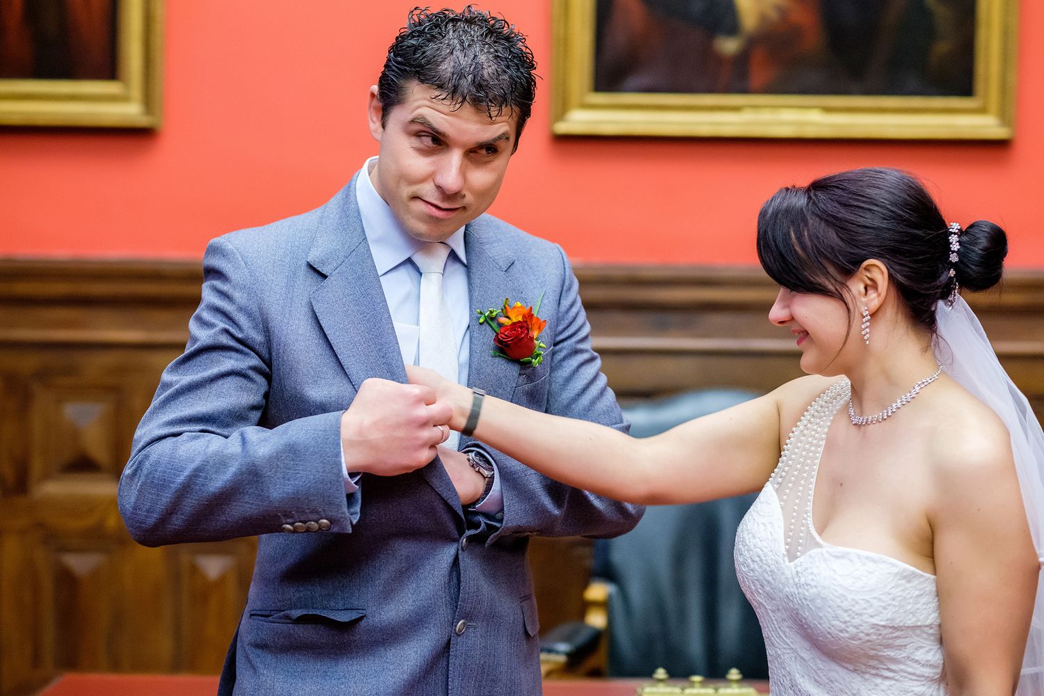 Couple exchanging rings during an elegant indoor wedding ceremony against a red wall with paintings.