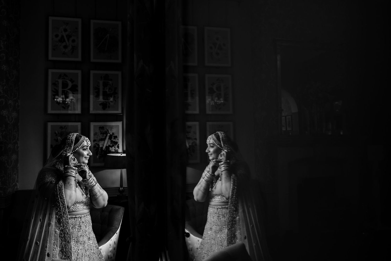 Artistic black and white reflection of a bride in traditional dress looking in a mirror with framed artwork on the wall.