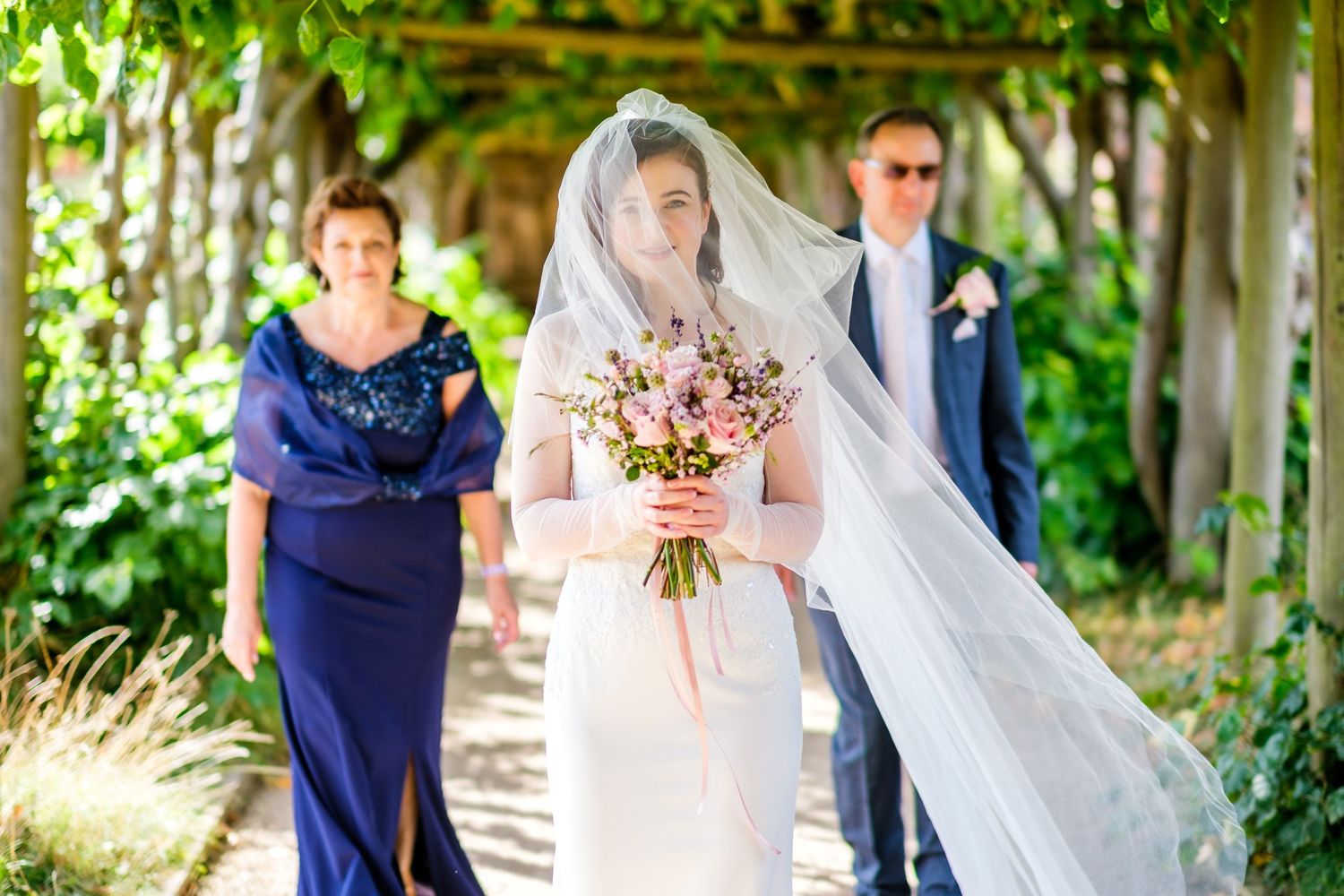 Outdoor wedding procession under a tree-lined path with natural greenery and sunlight.