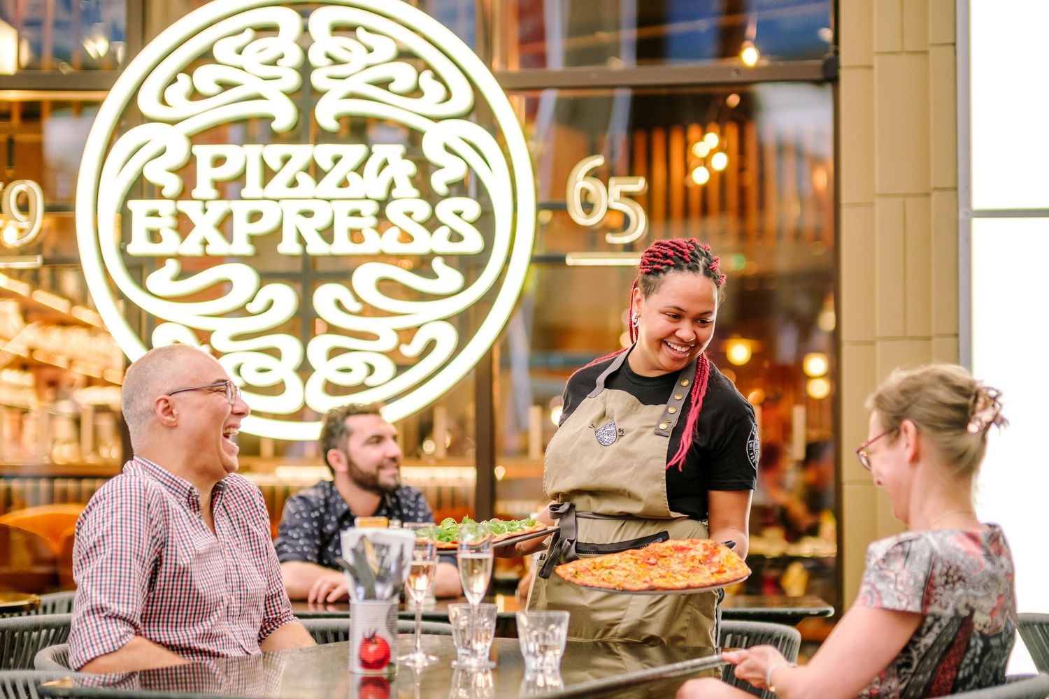 Illuminated Pizza Express logo and storefront with customers dining at outdoor tables.