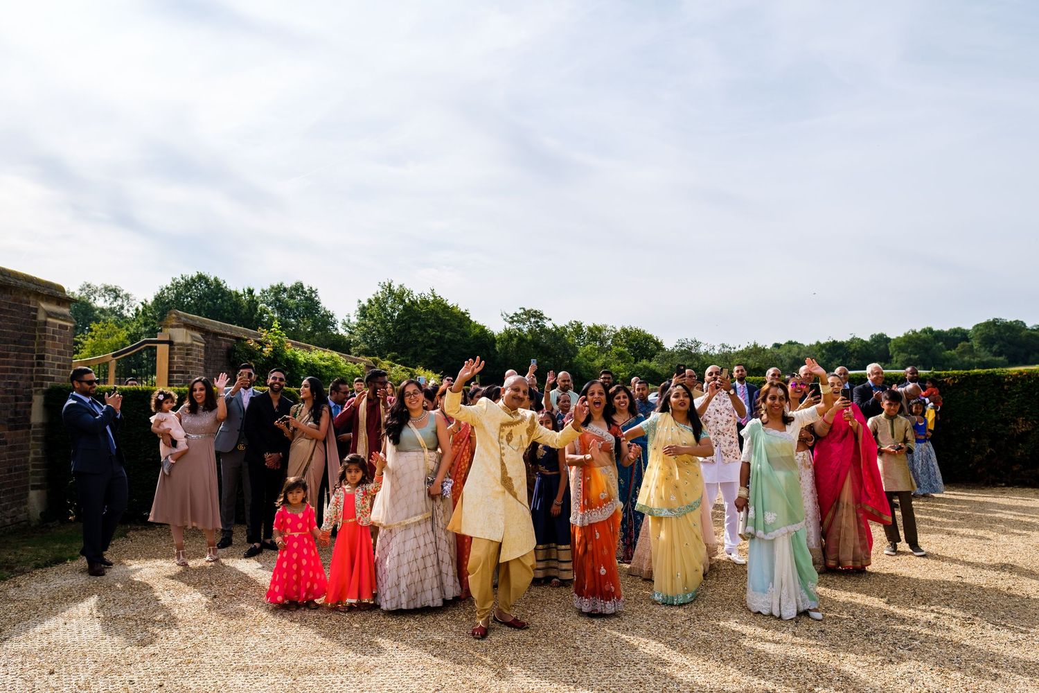 Large group of wedding guests in colorful traditional attire pose together outdoors for a group photo.
