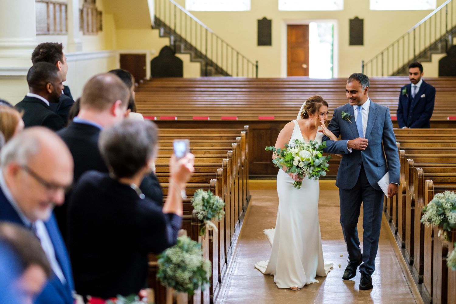 Newlyweds walk down the church aisle after ceremony while guests capture the moment on their phones.