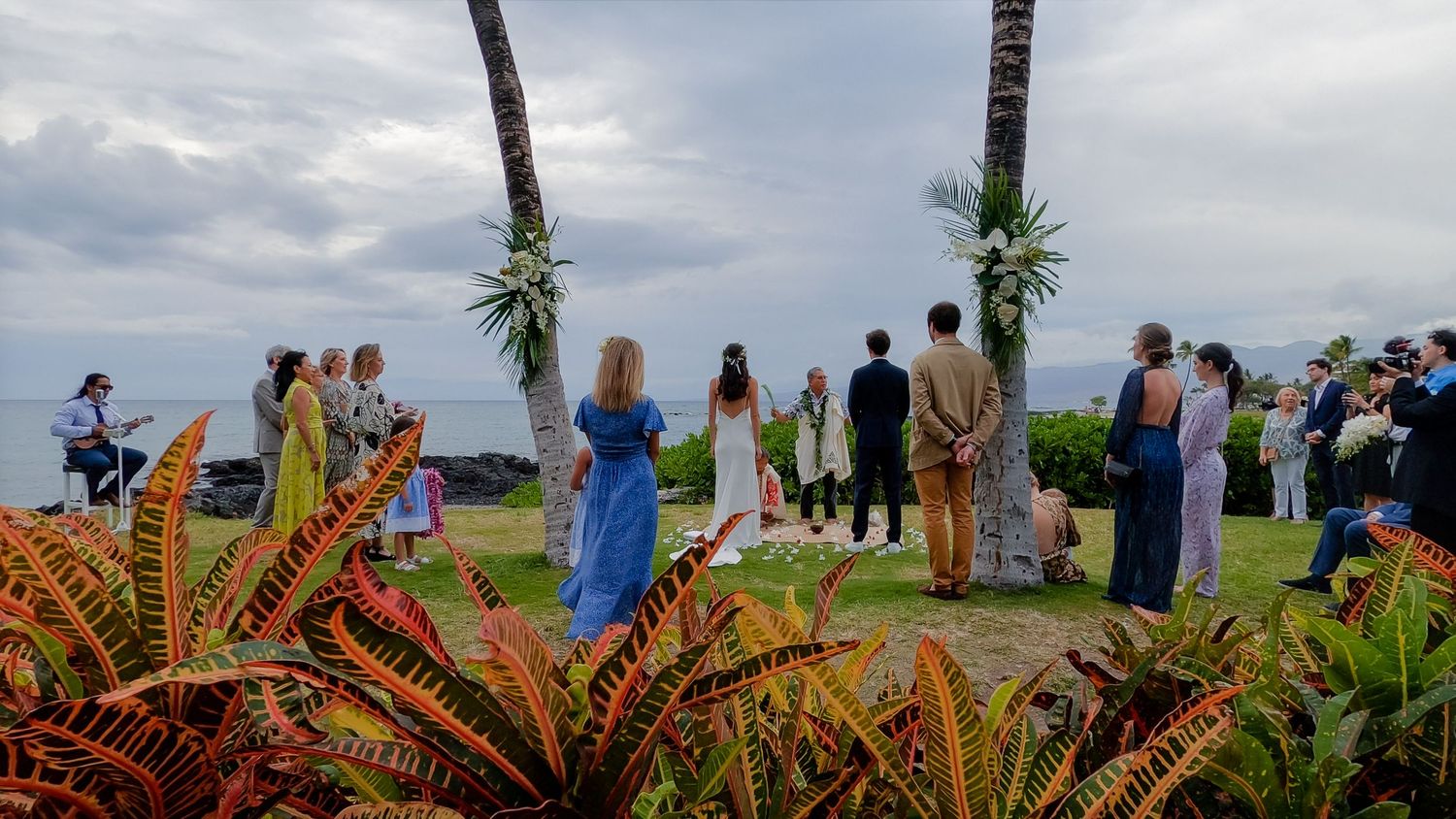 Wedding guests gather at a tropical oceanside ceremony with palm trees and colorful plants.