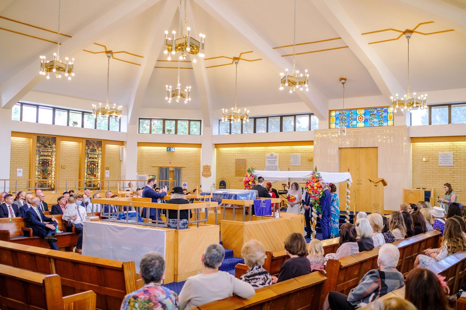 Bright synagogue interior with chandeliers and wooden pews filled with wedding guests during ceremony.