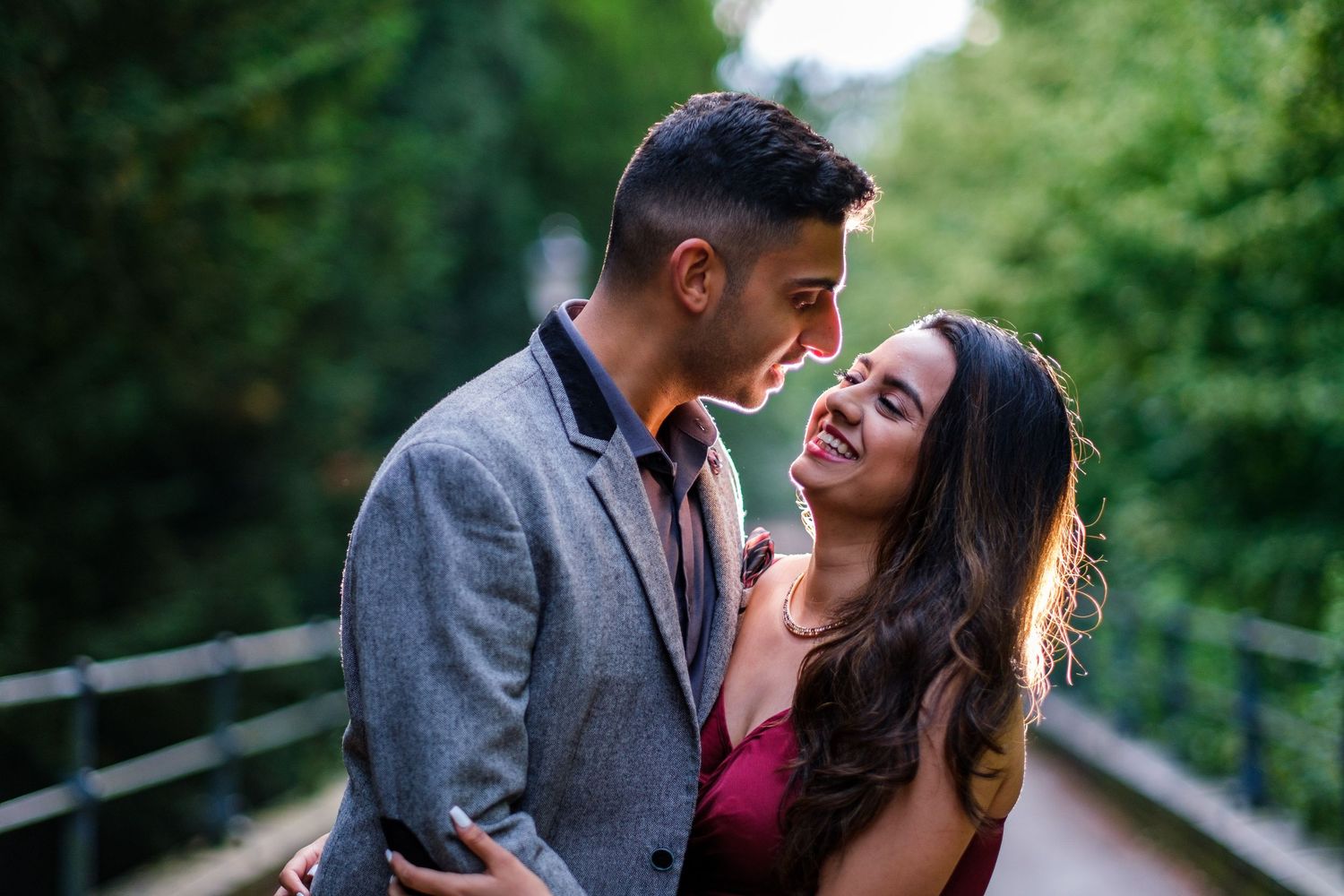 Romantic couple embraces and laughs together on a wooden bridge surrounded by lush green trees in summer.