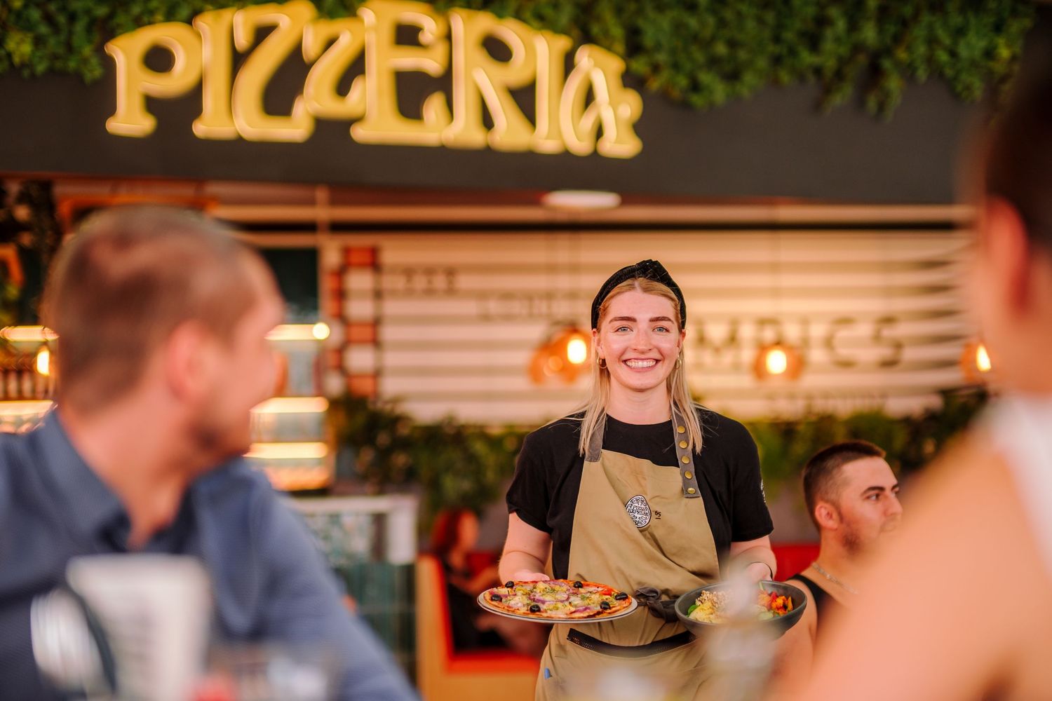 A server in a tan apron carries plates at an outdoor pizzeria with illuminated signage in the background.