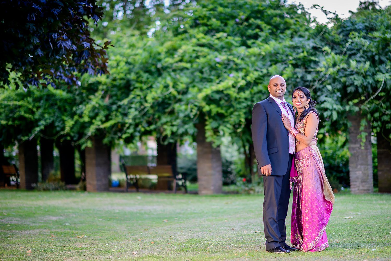 A couple poses outdoors in formal attire with a pink gown against a green garden background.