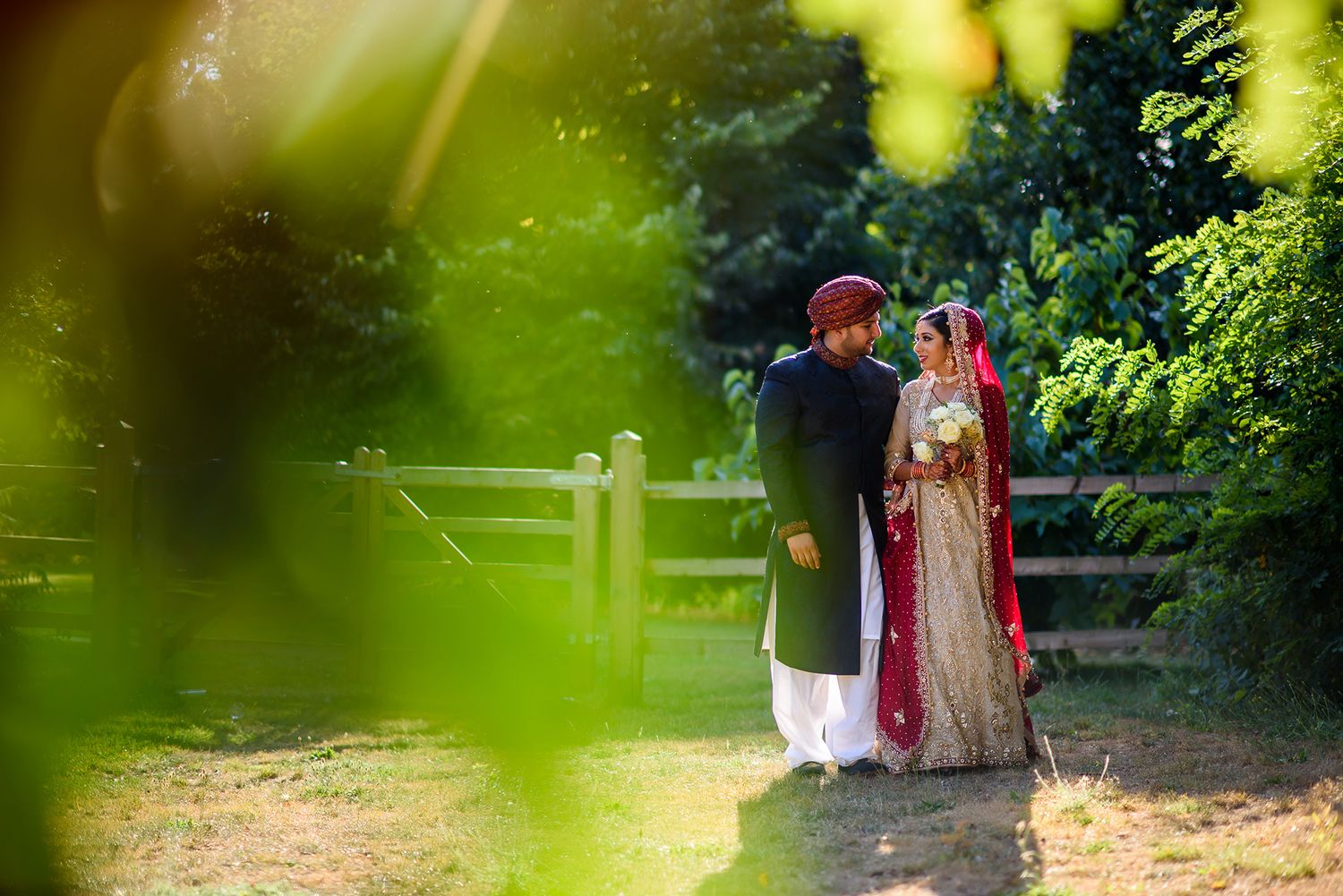 Artistic wedding photo through greenery showing couple in traditional South Asian attire.