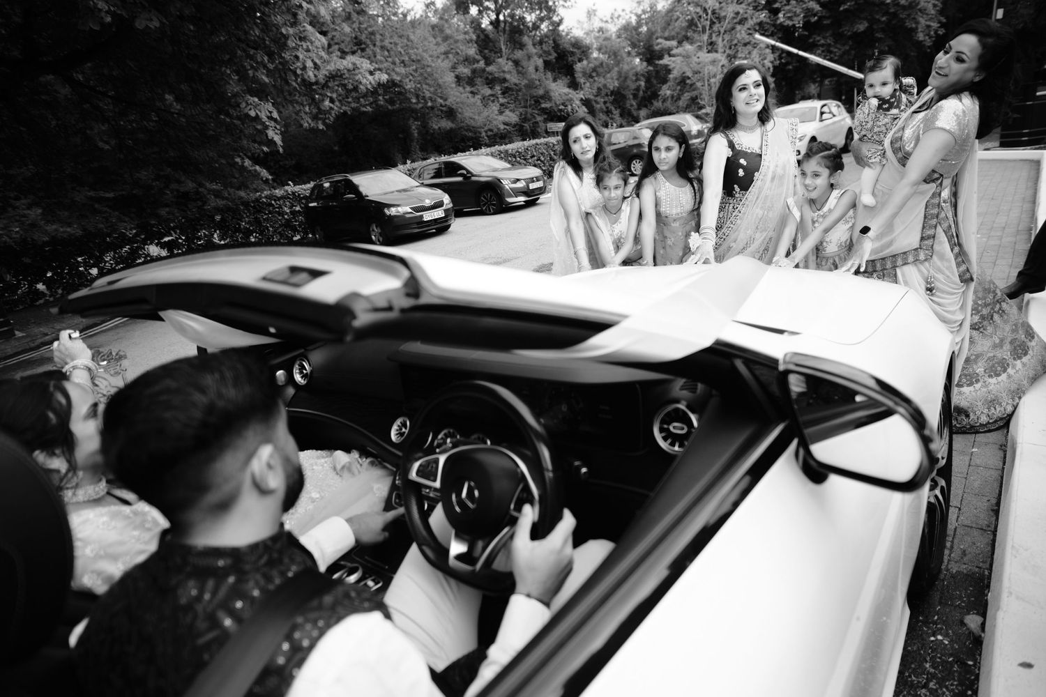 A black and white photo shows people gathered around a convertible car during a wedding celebration.