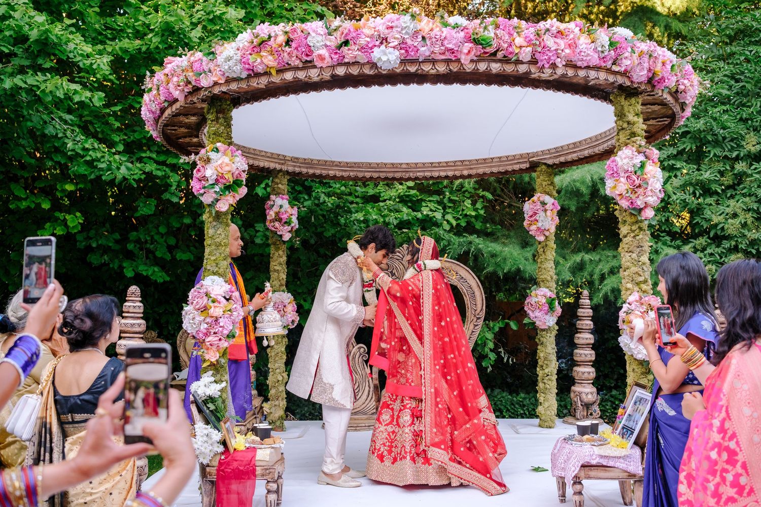 A couple stands beneath an ornate floral mandap decorated with pink blooms during an outdoor Indian wedding ceremony.