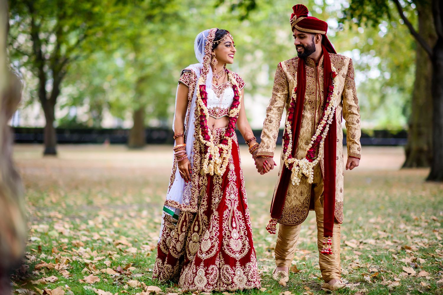 A couple in traditional Indian wedding attire walks through a scenic park during autumn.
