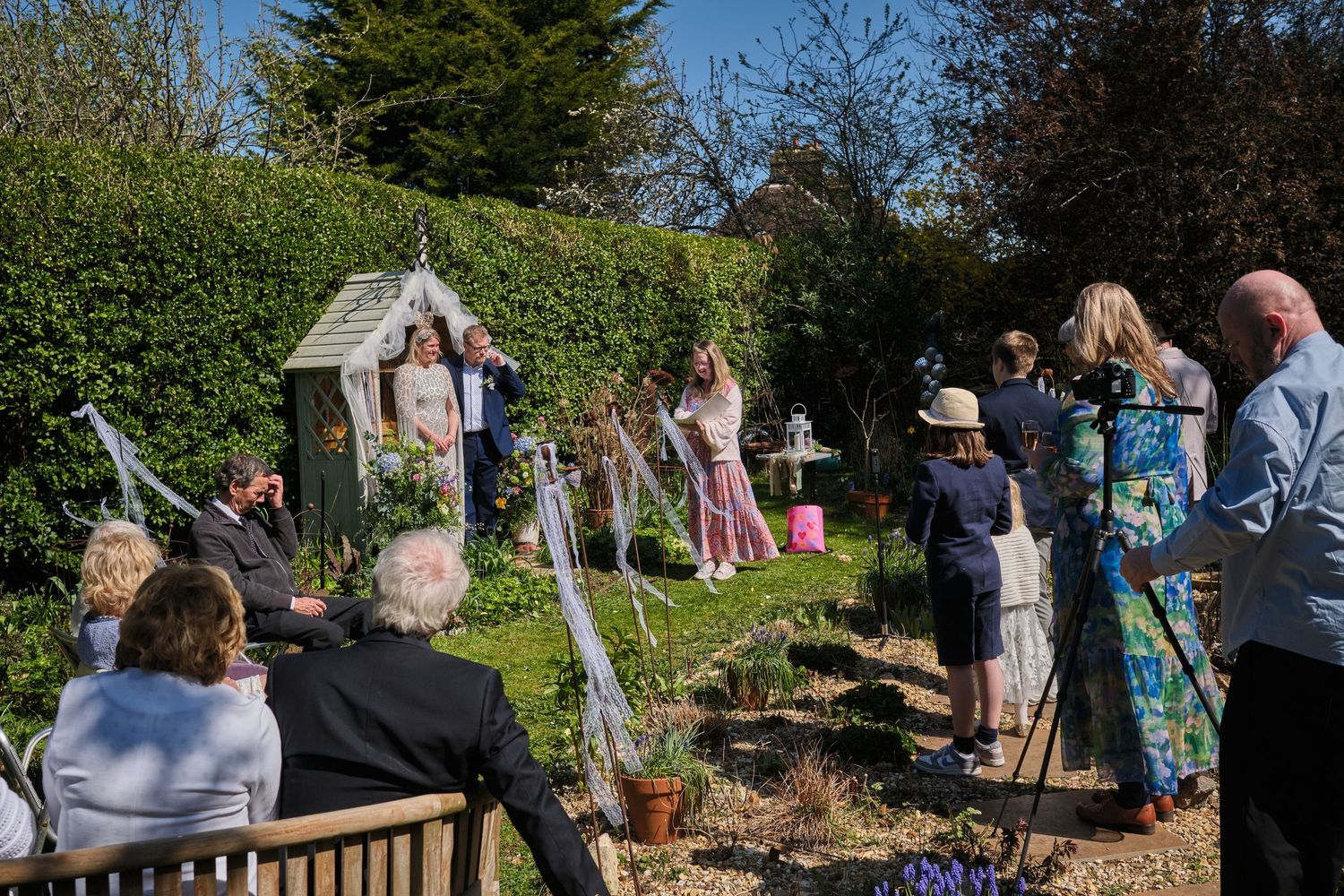 Garden wedding celebration with guests gathered among green hedges and trees.