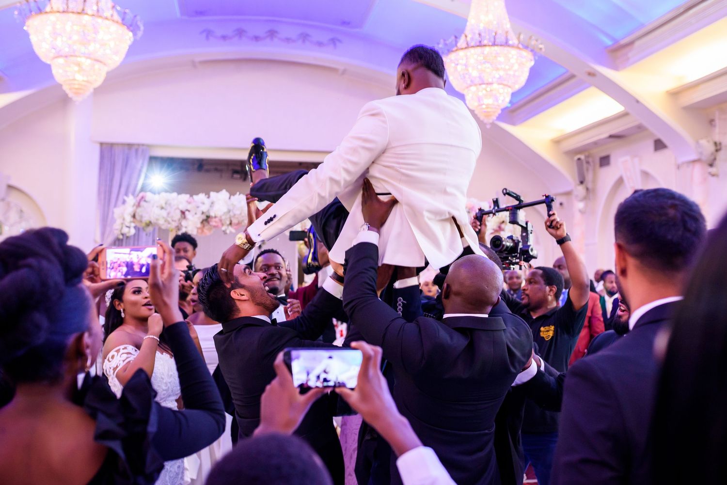 Elegant ballroom wedding celebration with crystal chandeliers and guests dancing in formal wear.