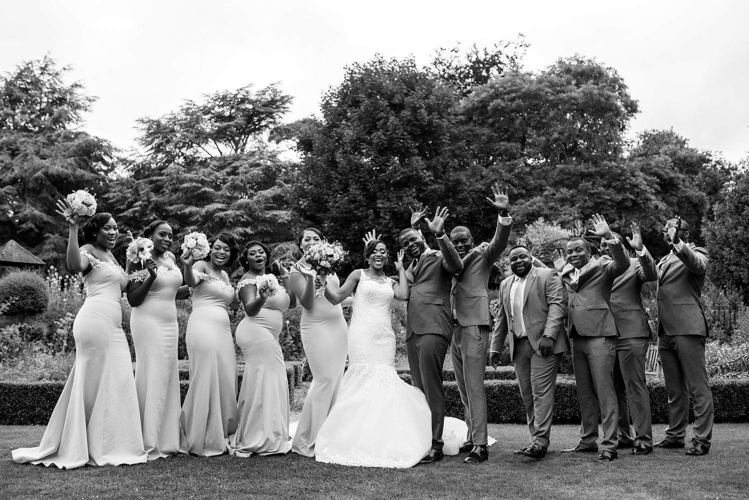 Wedding party members in formal attire pose together in a line outdoors with trees in the background.