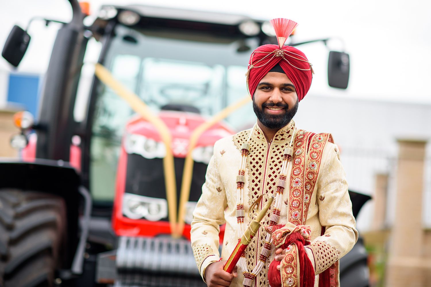 Groom in traditional Indian wedding attire standing in front of a red tractor.