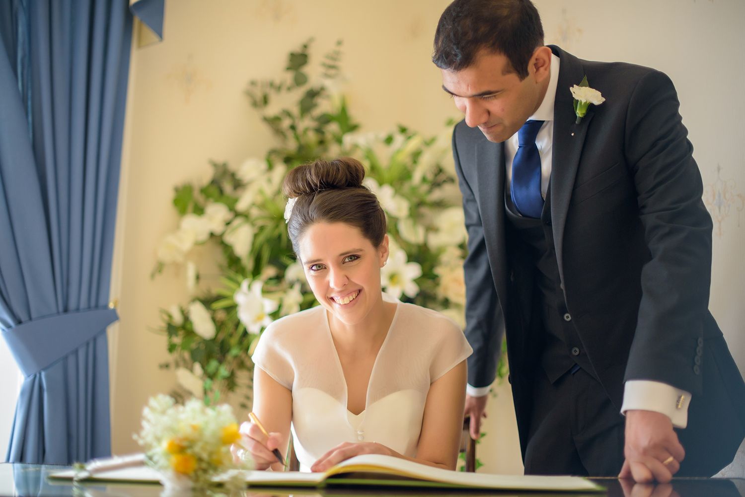 Couple signs marriage documents during their wedding ceremony in a room with blue curtains.