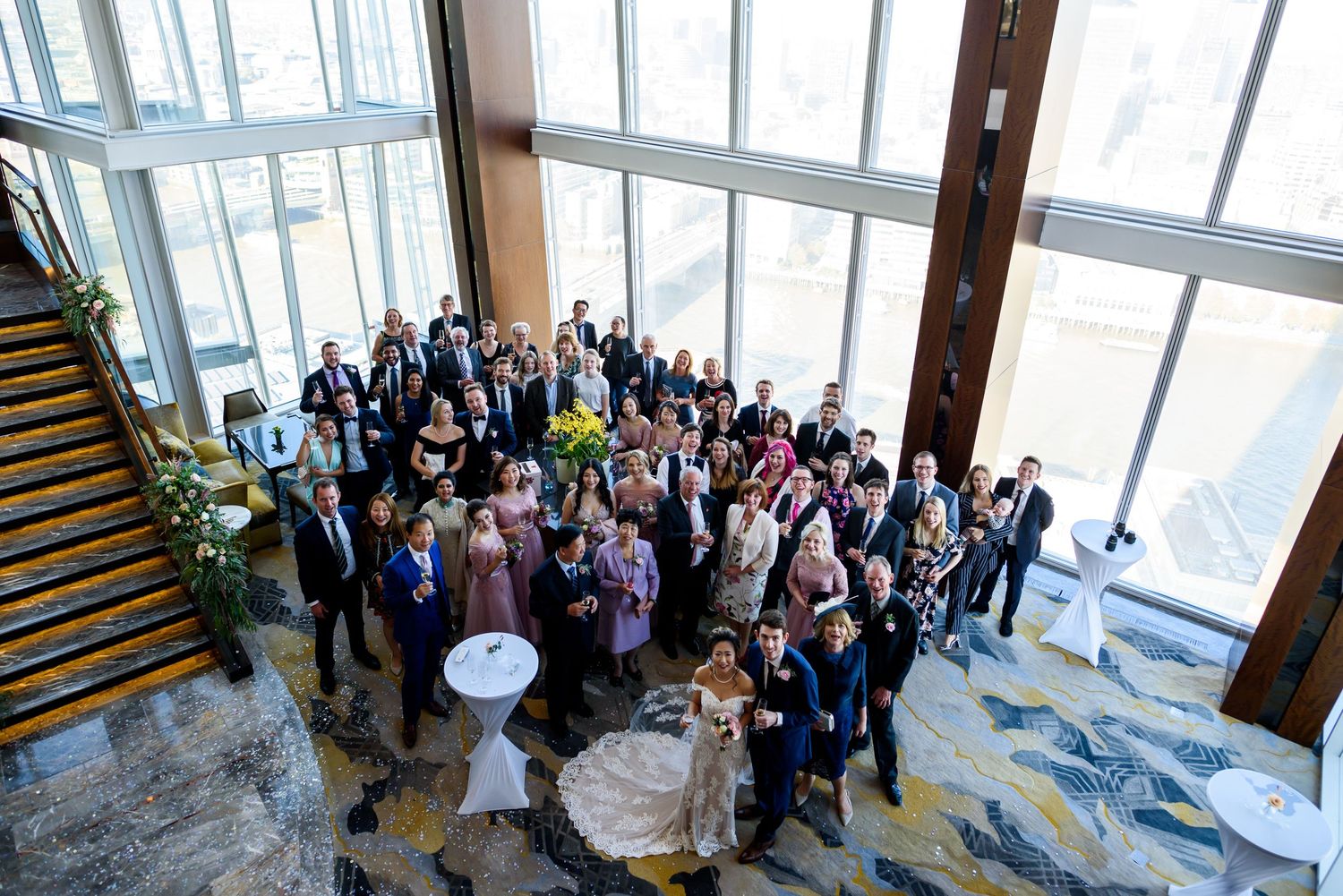 Large group of wedding guests gathered in a modern venue with floor-to-ceiling windows.