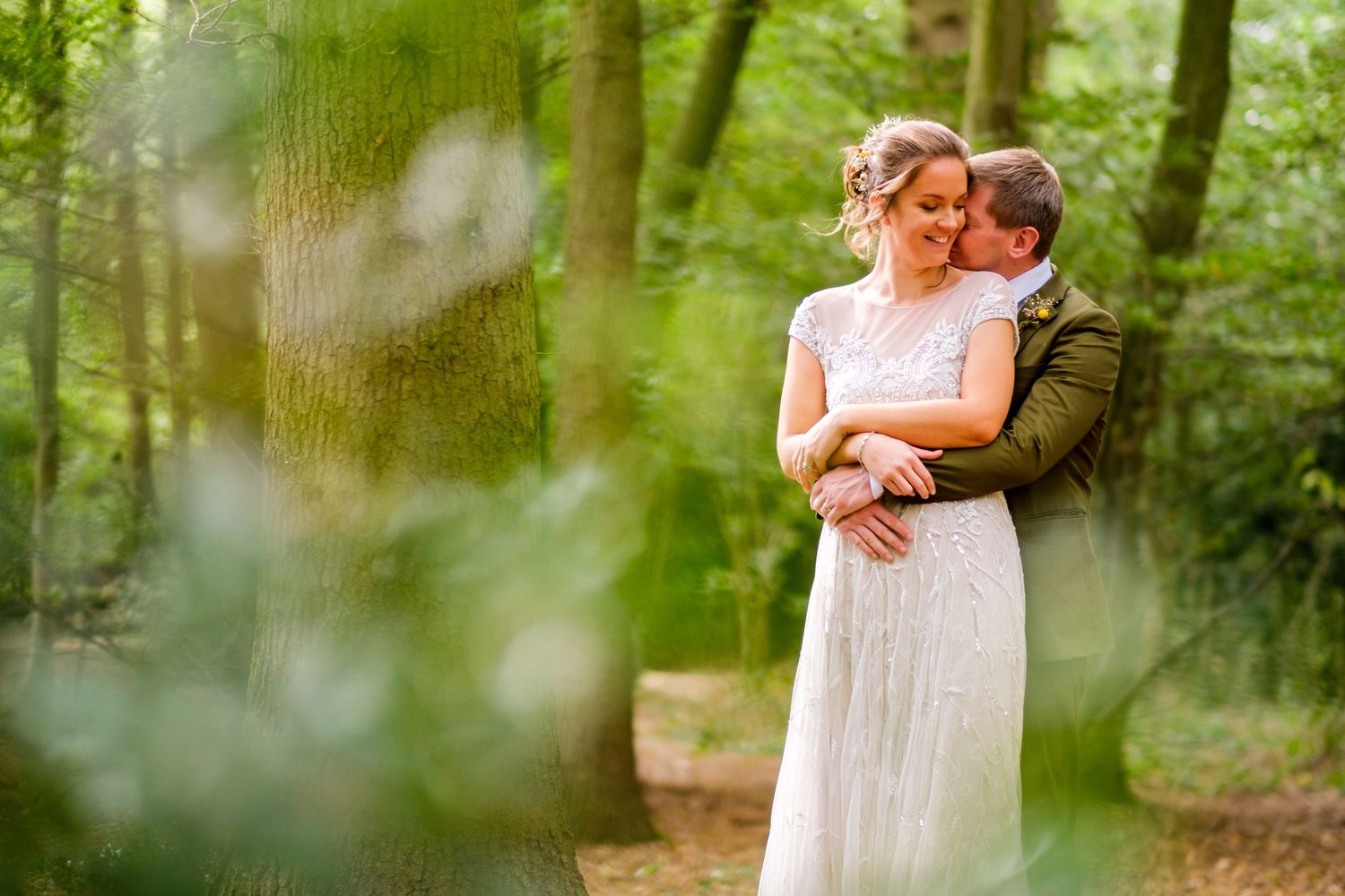 A romantic wedding couple embraces in a dreamy forest setting with soft natural lighting.