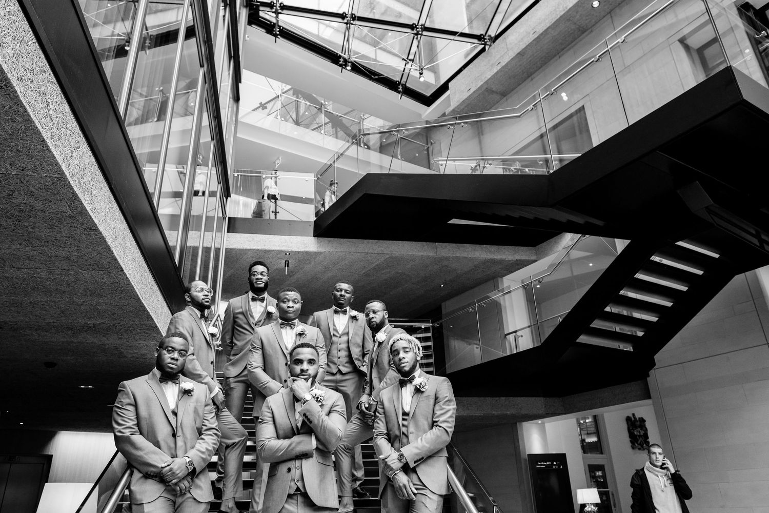 A group in formal attire poses together on a modern staircase in a building with glass ceilings.