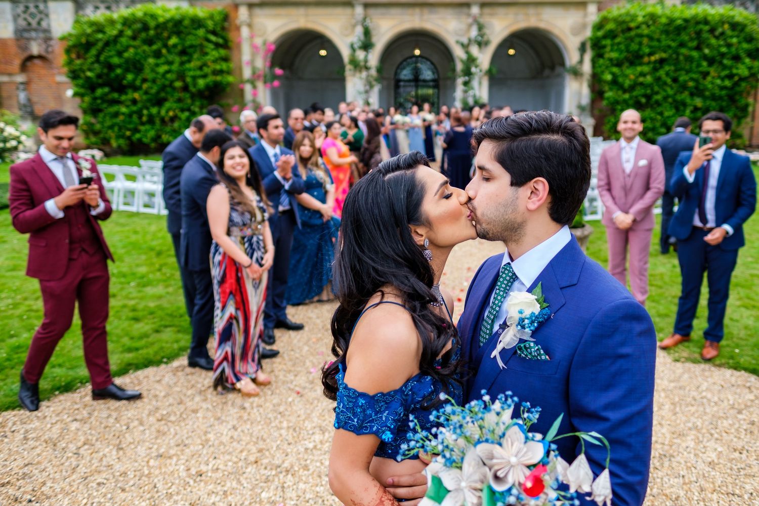 Wedding celebration on mansion grounds with guests gathered near ivy-covered archways.
