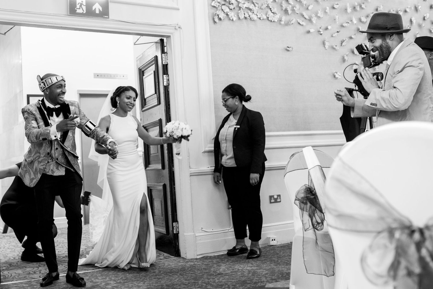 Wedding photographers capture candid moments during an indoor reception with white walls.