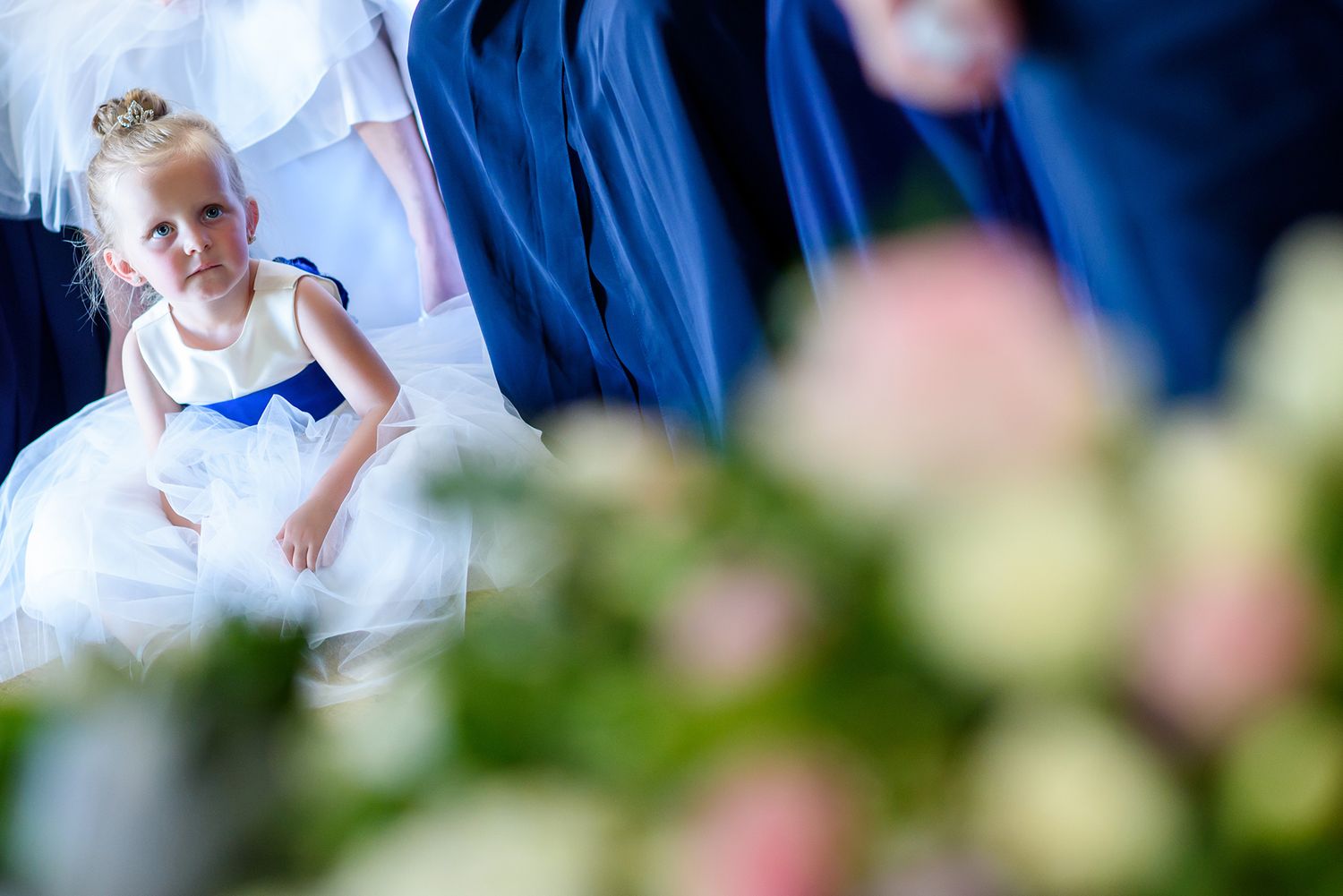A child in white formal attire seen through blurred flowers and blue fabric.
