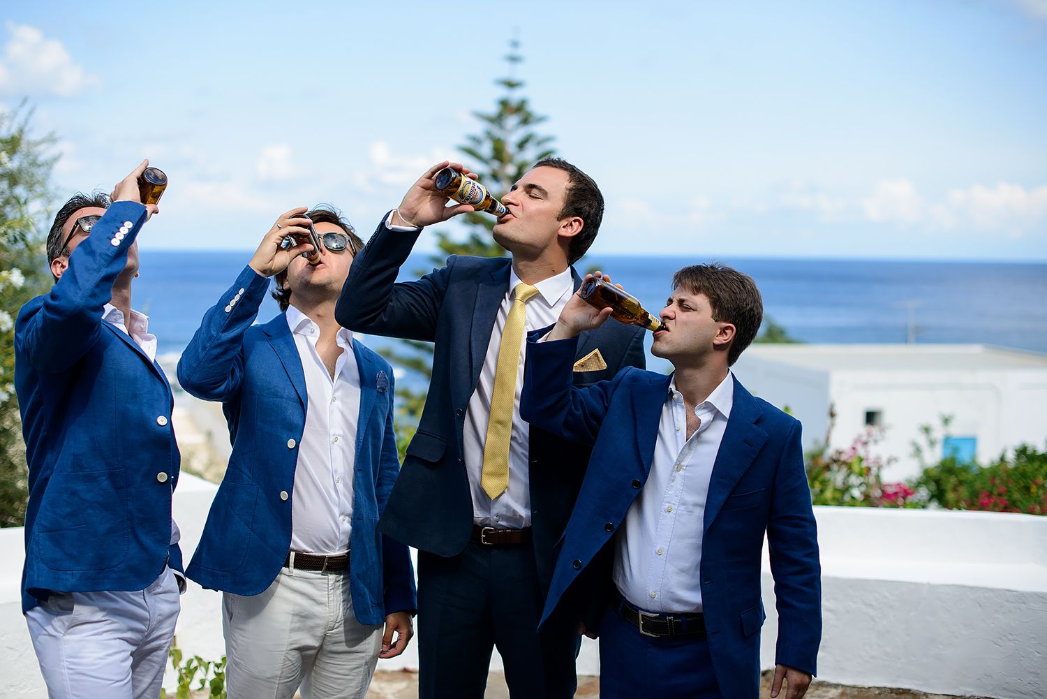 Groomsmen in navy suits toasting and celebrating outdoors by the ocean.