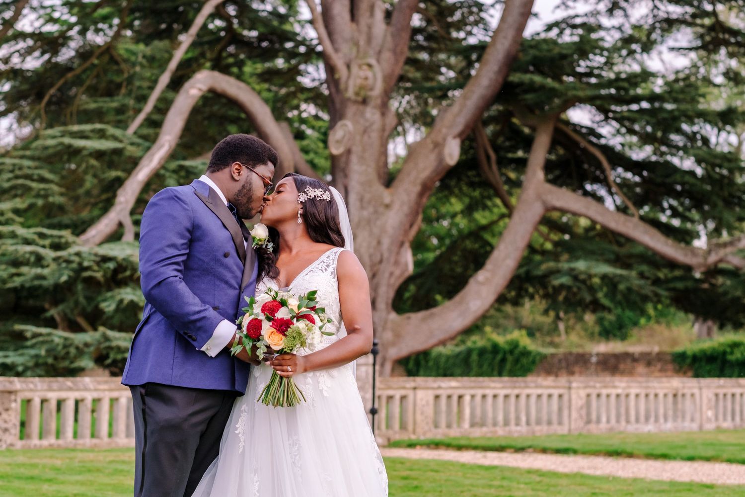 Newlyweds pose together under large trees in an outdoor garden setting.