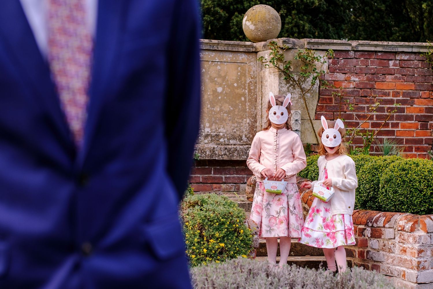 Two small children walk together in pink dresses and bunny costumes near a brick wall with greenery.