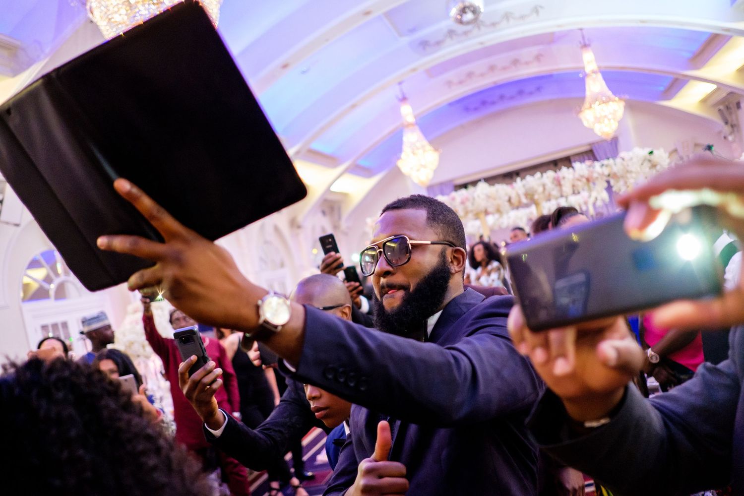 Chandelier-lit ballroom filled with celebrating wedding guests dancing with hands in the air.