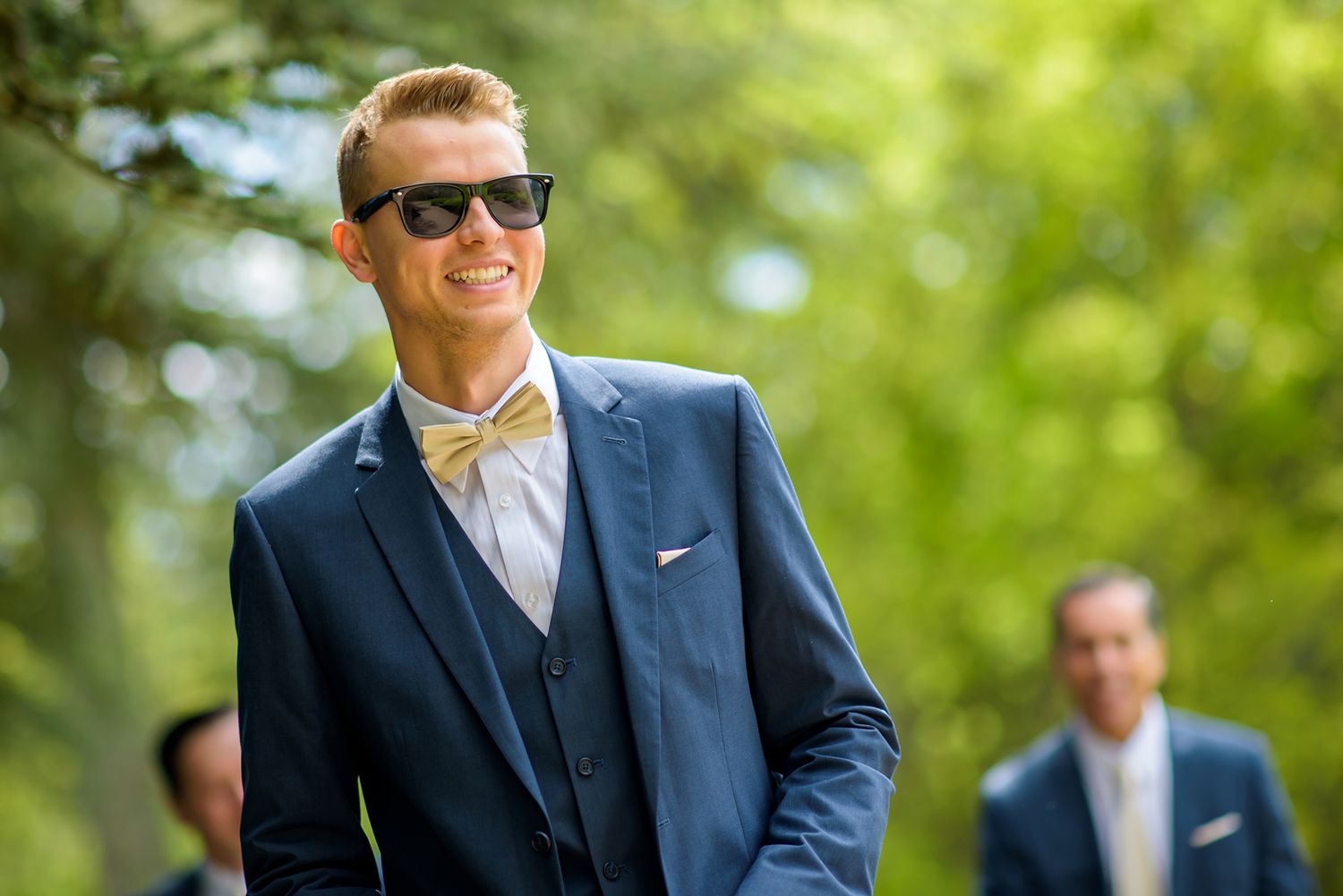 Groomsmen in navy suits and sunglasses stand together in a sunny outdoor setting.