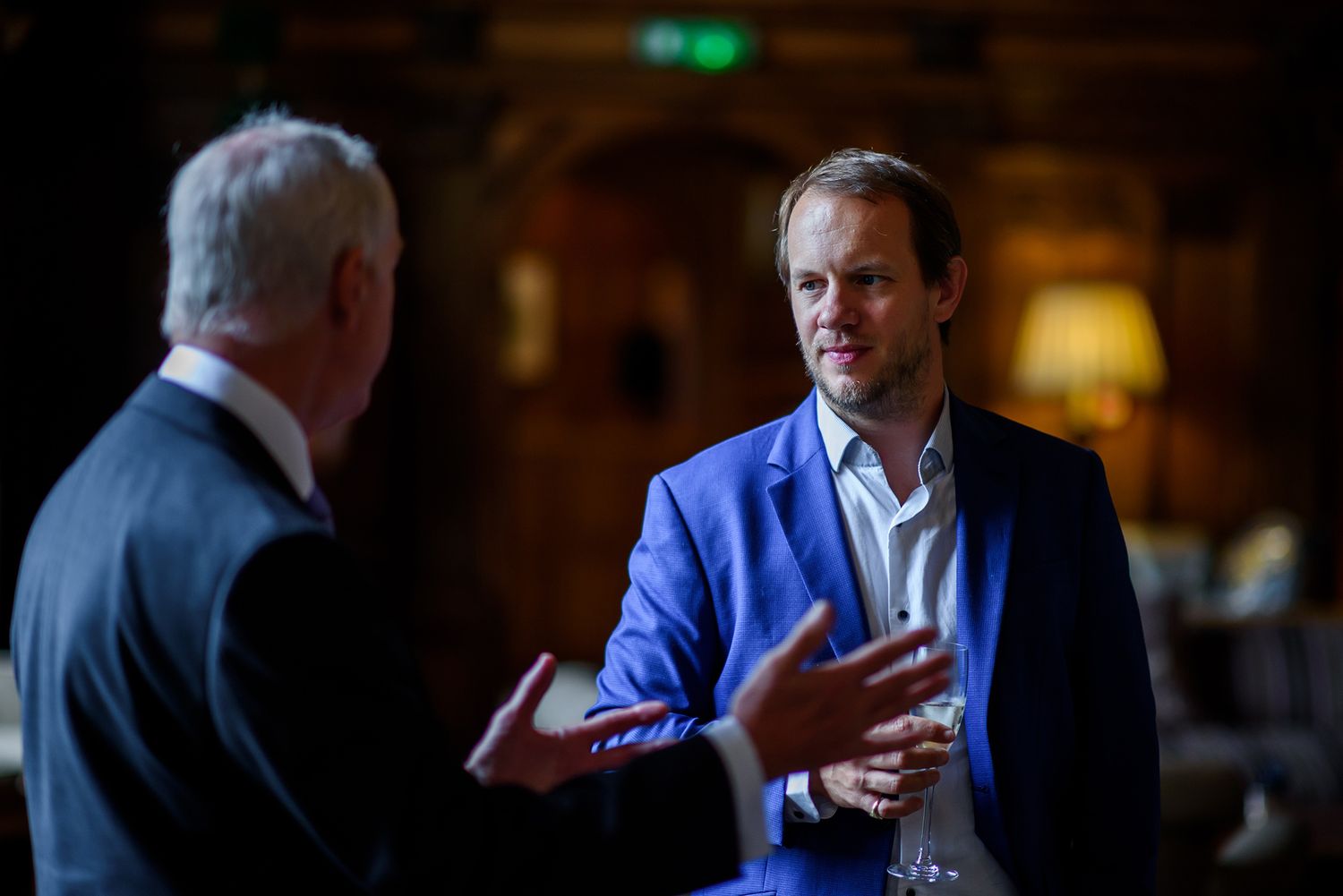 Two men engaged in serious conversation in dimly lit formal setting.