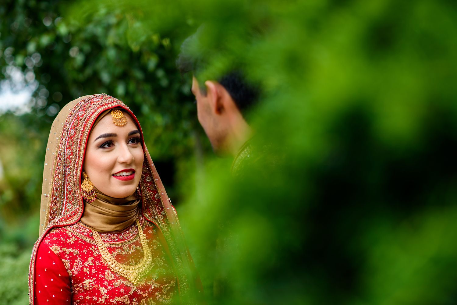 Beautiful traditional bride in red and gold attire poses against lush green outdoor background.