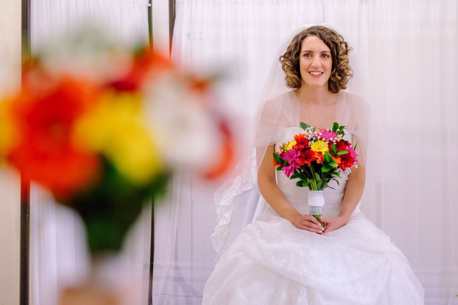 A bride in a white wedding dress holds a vibrant red and orange bouquet while standing against a light background.