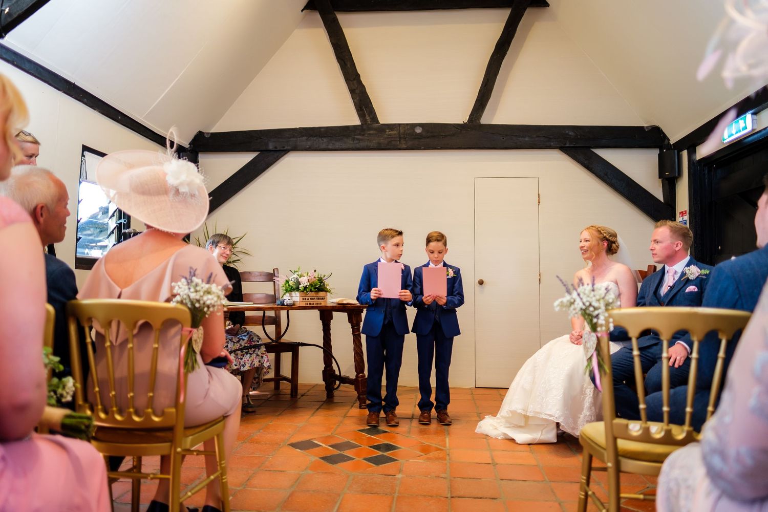 Wedding ceremony taking place in a rustic room with exposed wooden beams and guests seated in wooden chairs.