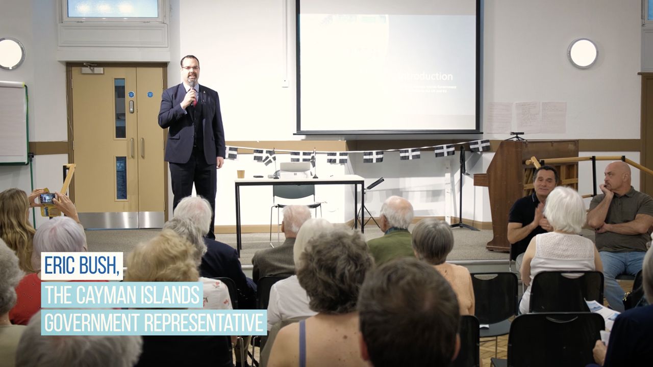 Eric Bush, Cayman Islands Government Representative, speaks at a public meeting with an audience seated before him.