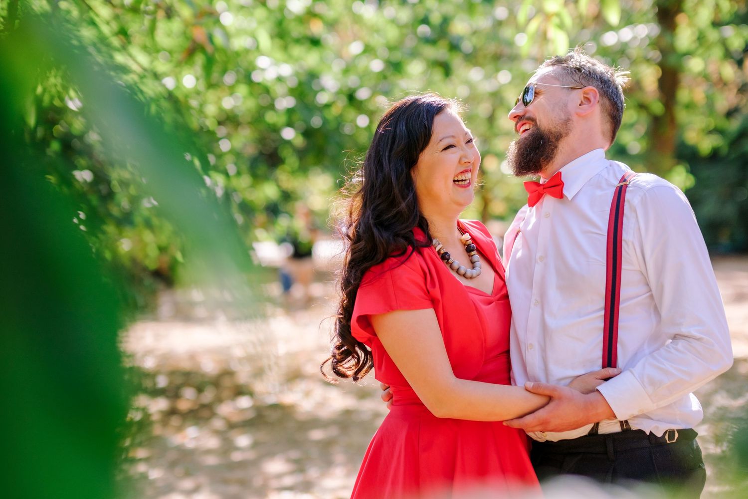 A couple in red and white attire share romantic moments while walking through a sunlit garden setting.