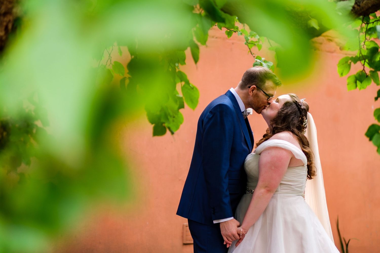 Couple sharing a romantic moment against a coral-colored wall with green foliage in the foreground.