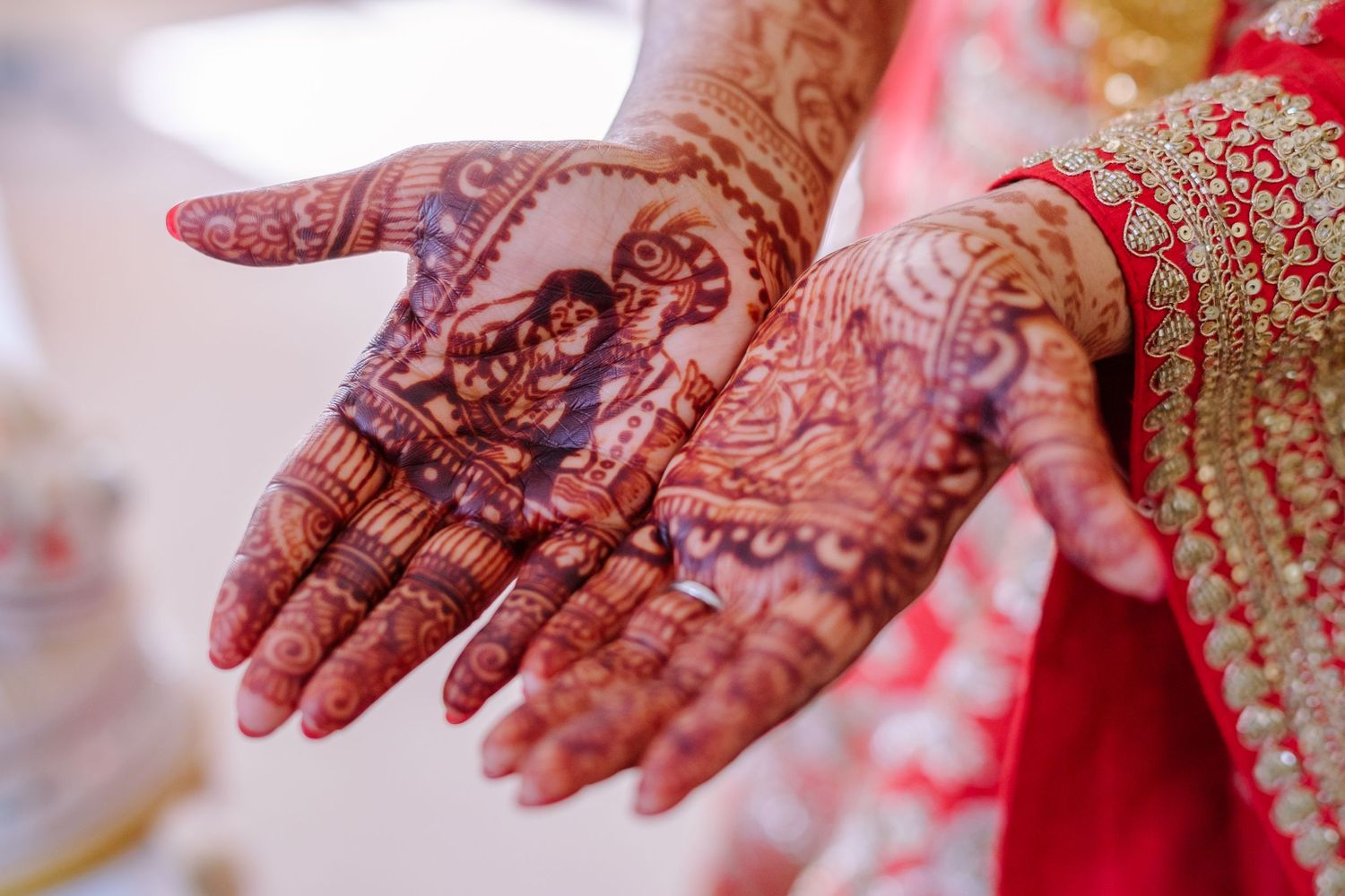 Intricate henna mehndi designs adorning hands against a red and pink traditional wedding backdrop.