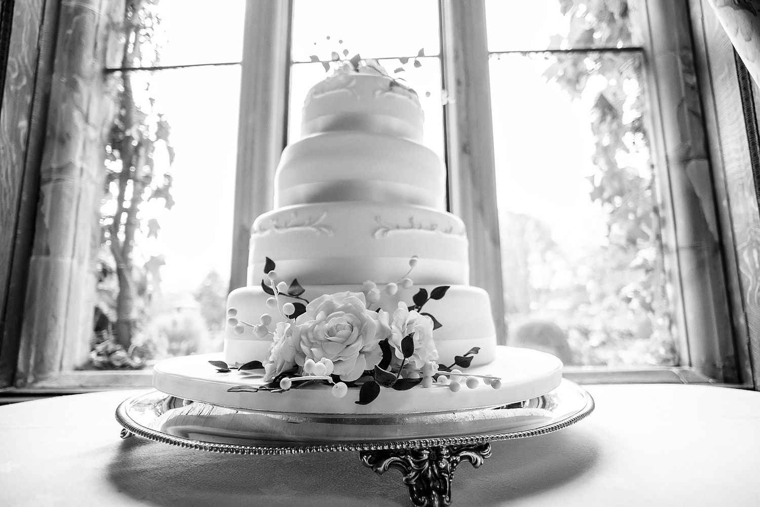 Wedding cake decorated with delicate white flowers and ribbon displayed in front of a bright window.