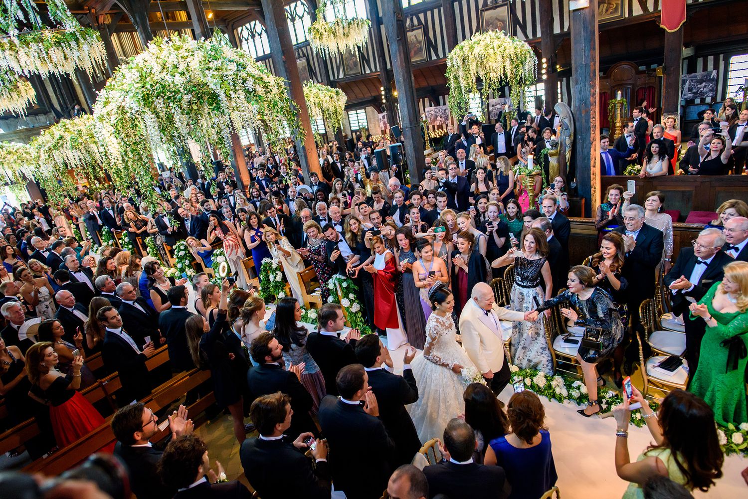 Large wedding reception crowd celebrating under ornate ceiling decorations and hanging greenery.