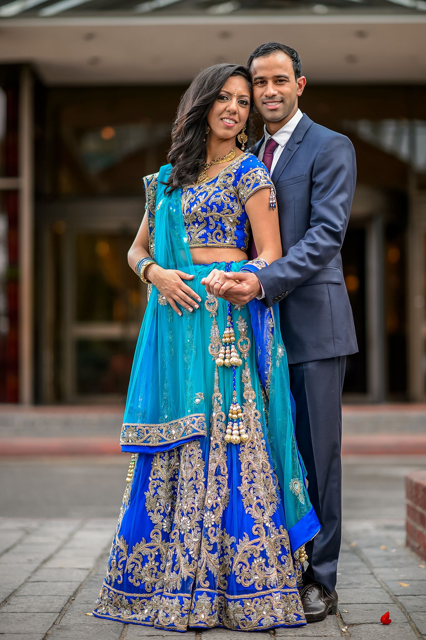 Couple in traditional Indian wedding attire poses outdoors, with bride wearing blue and gold lehenga.