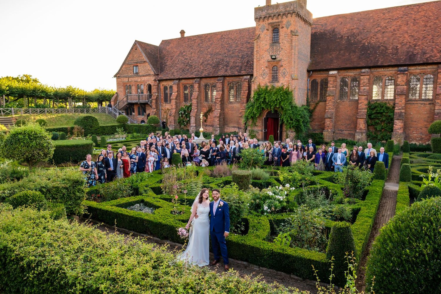 Aerial view of wedding guests gathered in formal garden with historic brick building backdrop.